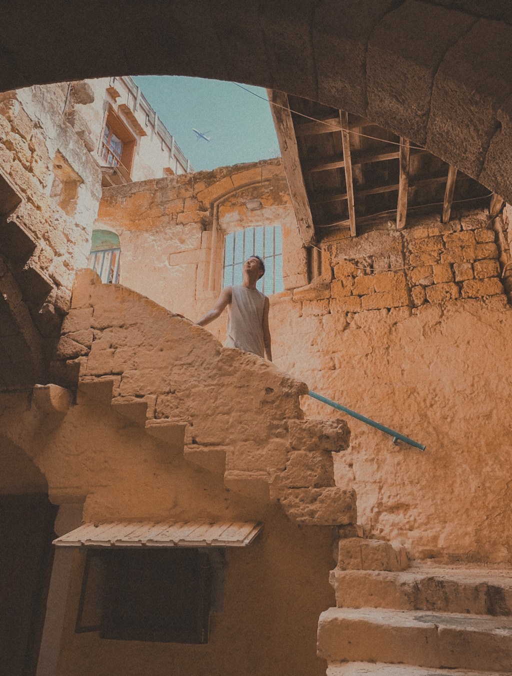 A man stands in the centre of frame on old medieval stairs. A blue sky has a plane flying above.