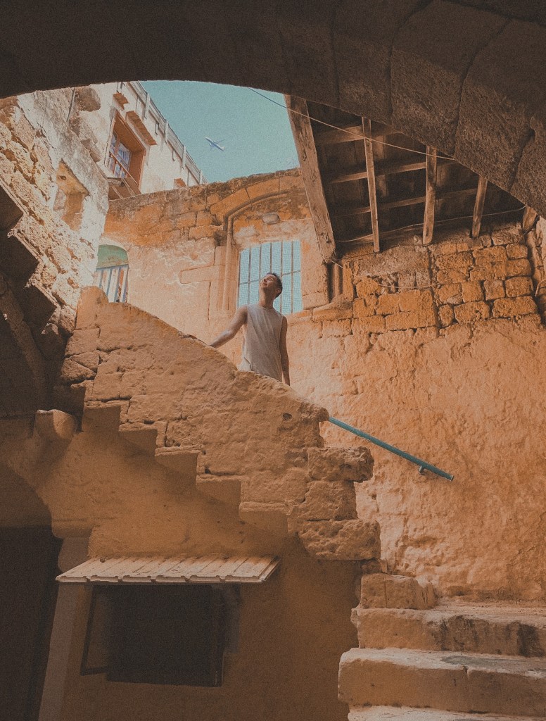 A man stands in the centre of frame on old medieval stairs. A blue sky has a plane flying above.