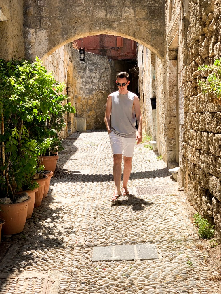A man in a grey vest walks down a cobbled street of Old Town Rhodes